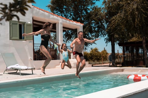 Parents and daughter holding hands, joyfully jumping simultaneously into a swimming pool under a bright blue sky