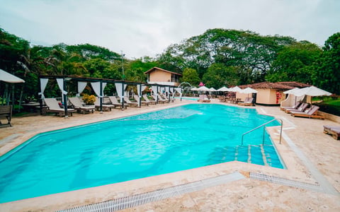 Resort swimming pool with turquoise water, white canopy loungers, and lush green trees in background
