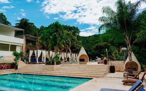 Luxury resort with turquoise swimming pool, white buildings, tropical palm trees, and mountain backdrop under blue sky