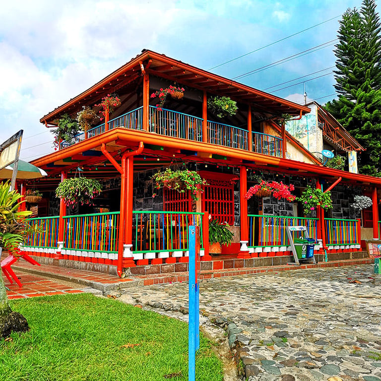 Red wooden house with two-story architecture, wraparound porches, and flowering plants, surrounded by manicured lawn and stone pathway