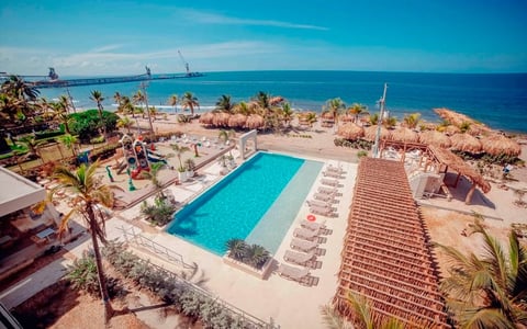 Aerial view of a beachfront resort with turquoise swimming pool, loungers, palm trees, and Mediterranean sea under clear blue sky